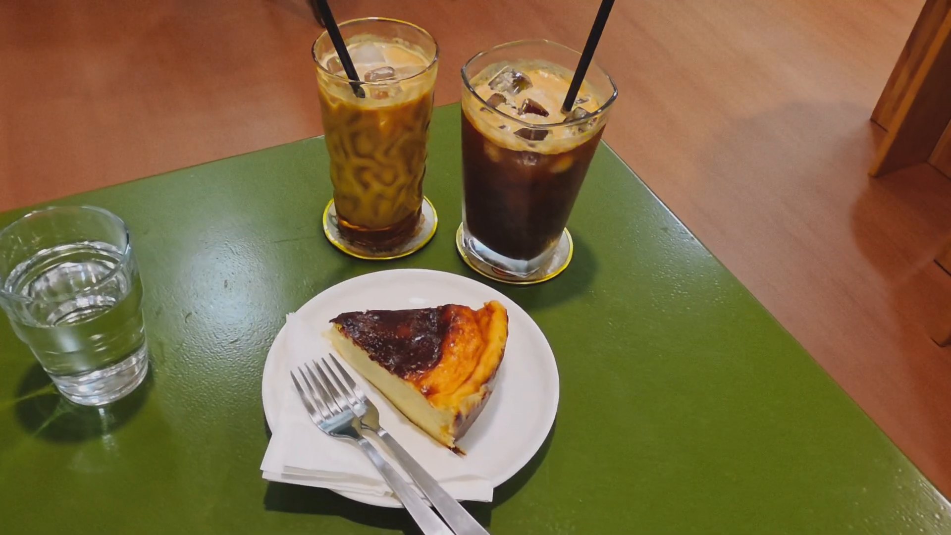 An iced white coffee and an espresso tonic beside a slice of burnt cheesecake on a green café table.