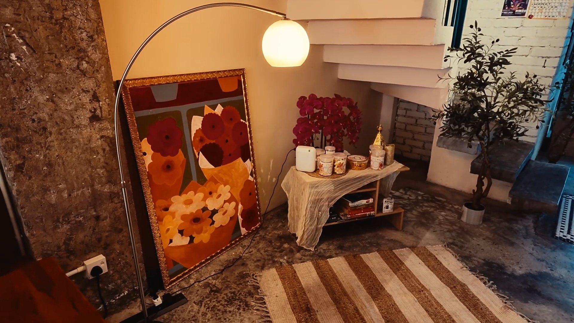 A quiet café corner with a floor lamp, a flower painting, and a striped rug.